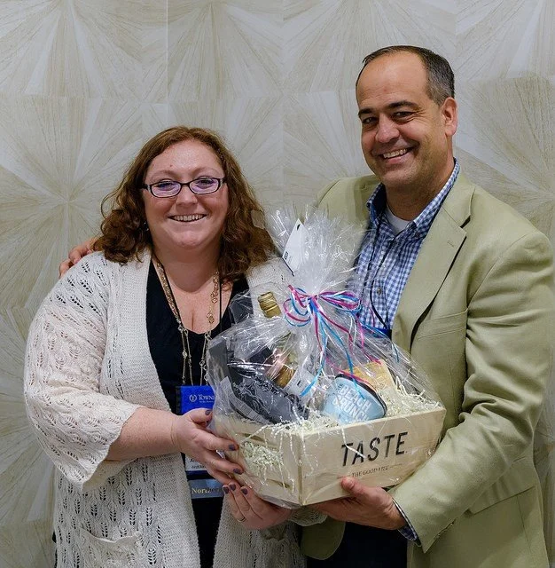 A smiling woman with glasses and curly hair wearing a white cardigan, standing next to a man in a beige blazer and blue checked shirt, holding a gift basket decorated with ribbon, against a tiled wall background.