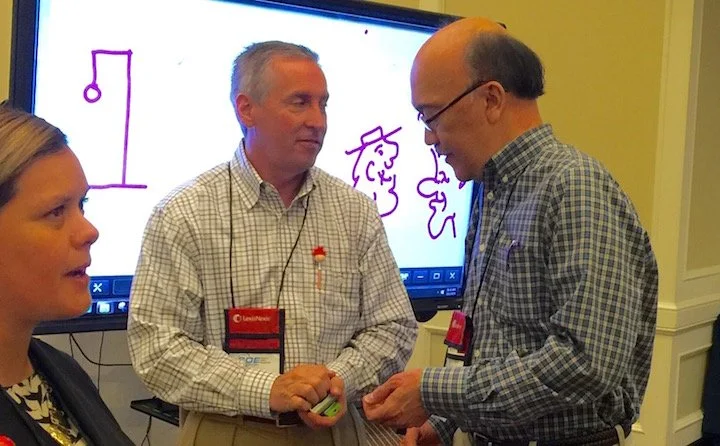 Three people engaged in conversation at a professional event, with a man and woman wearing name tags, in front of a screen with a digital drawing of cartoon faces.
