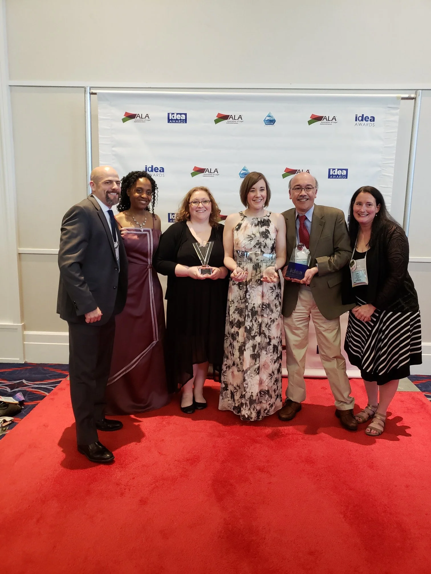 Group of six diverse people standing on a red carpet in front of a white backdrop with logos, holding awards.