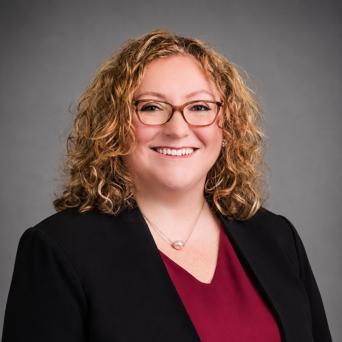 Professional headshot of a woman with curly blonde hair, glasses, and a friendly smile, wearing a black blazer and maroon top, against a gray background.