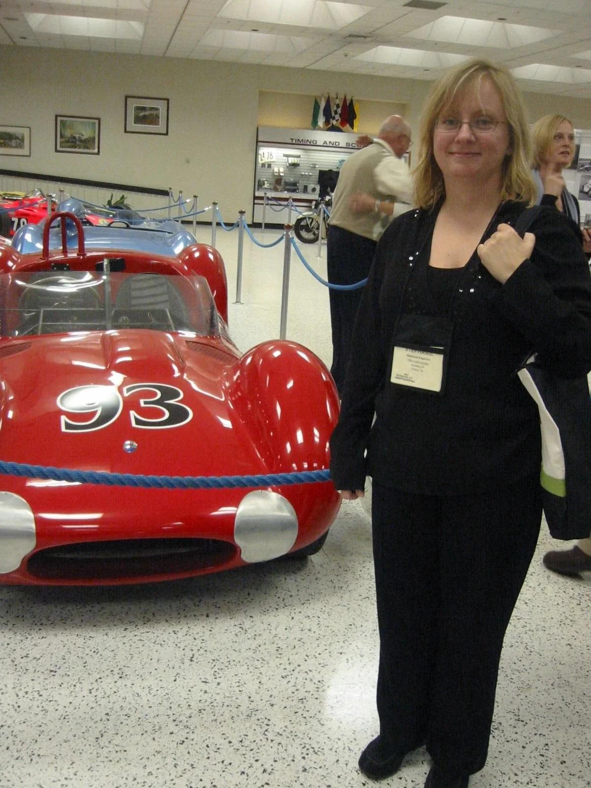 A woman with blonde hair and glasses standing in front of a red vintage race car with the number 93, inside a museum or exhibition hall.