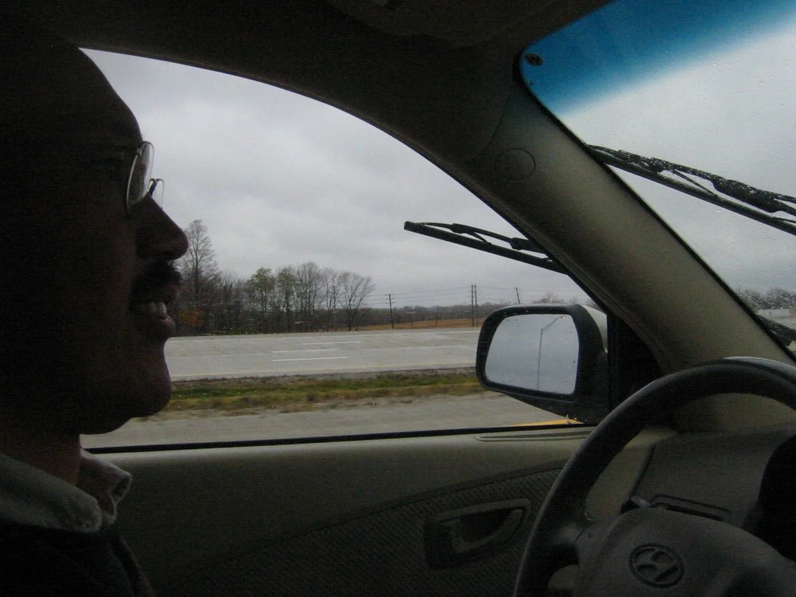 A person with glasses smiling inside a car, with a view of a rainy highway and overcast sky through the windshield.