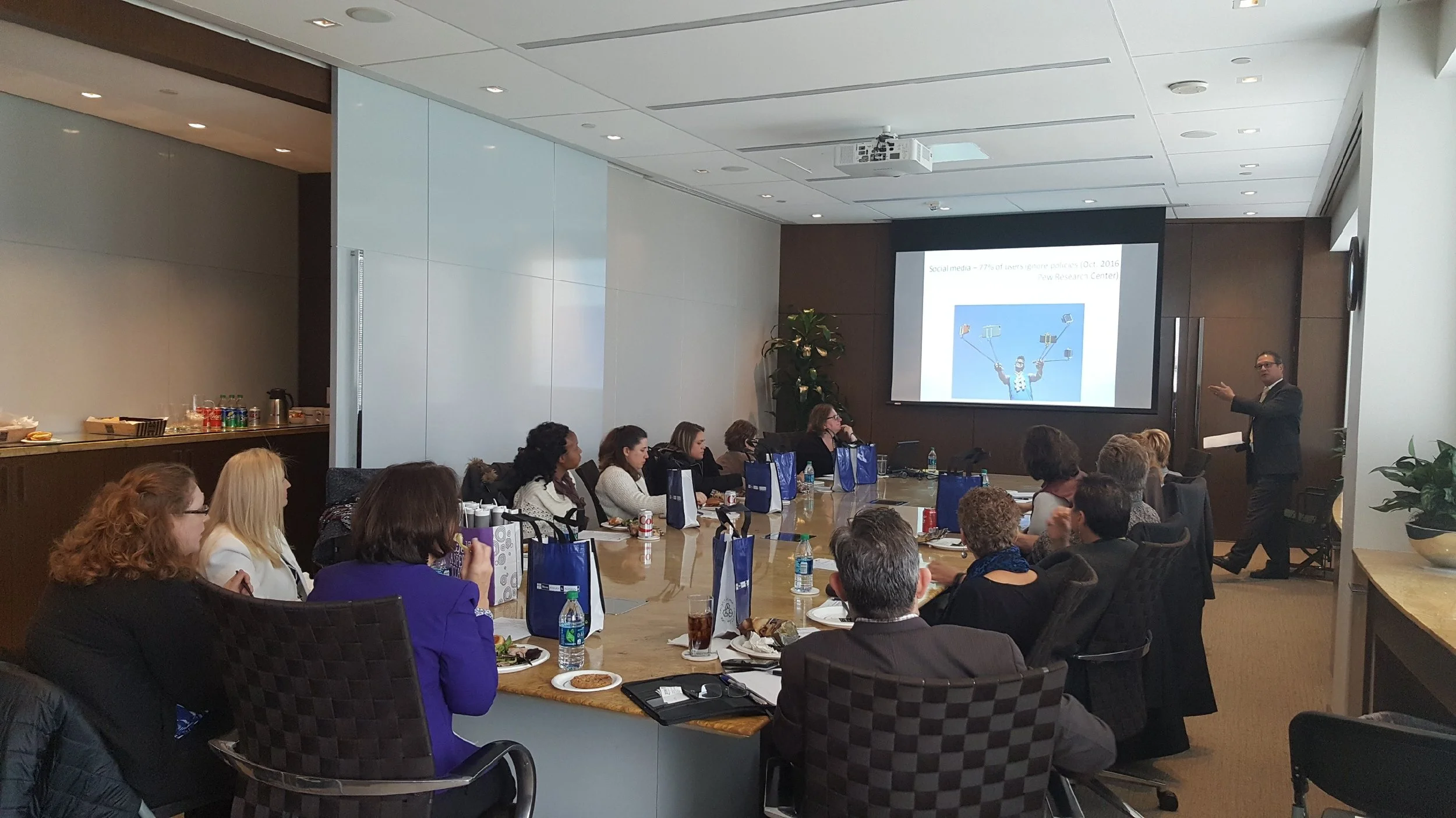 A conference room filled with people seated at a large table, listening to a presentation given by a man standing by a screen displaying a slide about social media.