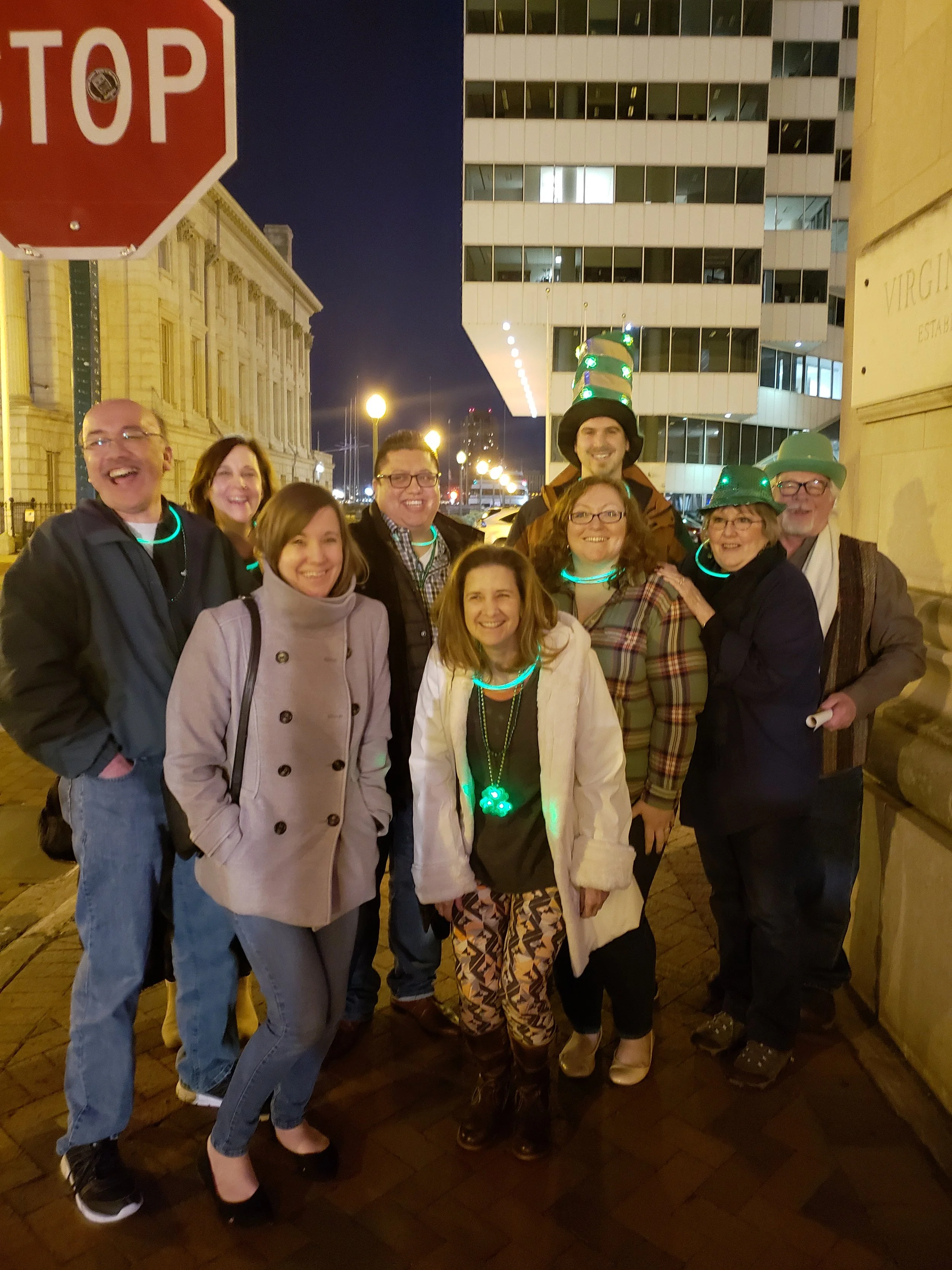 A group of ten people standing on a city sidewalk at night, smiling at the camera. Some are wearing glow-in-the-dark necklaces and hats, and one person is wearing a tall, green hat with lights. There is a stop sign in the upper left corner, and in th