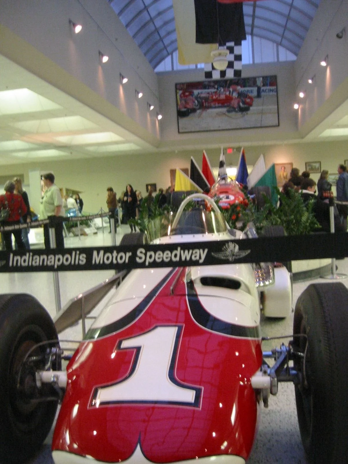 A racing car on display at the Indianapolis Motor Speedway, surrounded by a crowd of people. The car is white with red and black accents and features the number 3. In the background, there are flags and a large photograph of a racing scene.