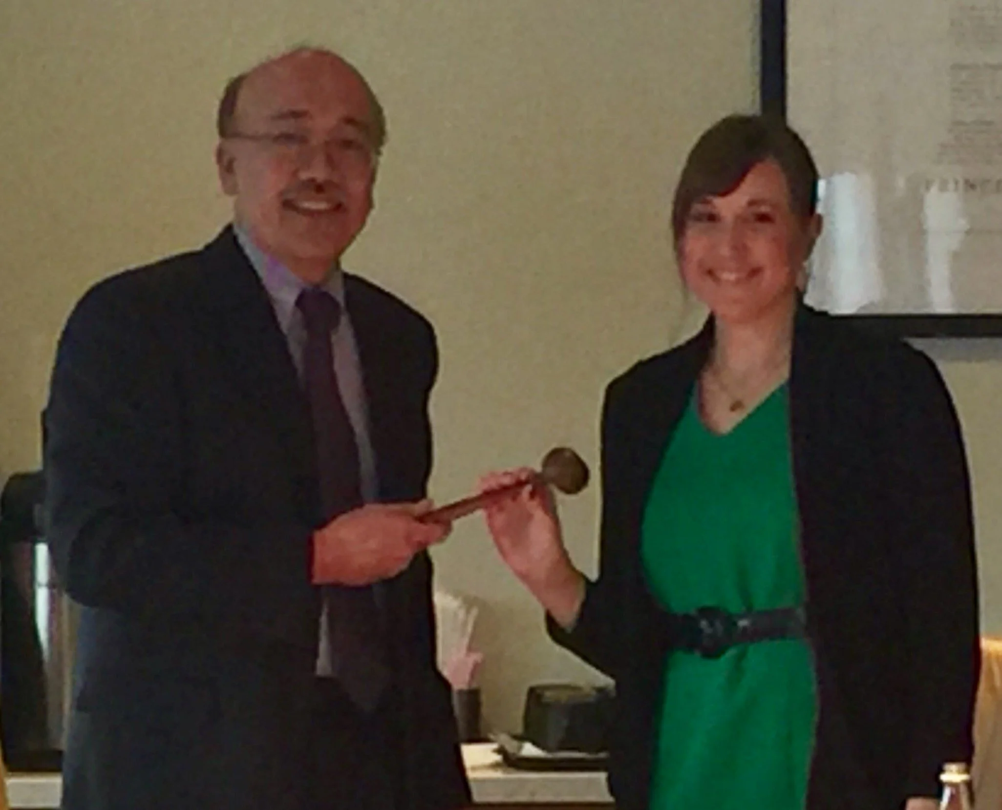 Two people, a man and a woman, standing side by side at an event. The man is wearing a dark suit and a tie, and the woman is dressed in a black blazer over a green top. They are smiling and the woman is holding an award or plaque.