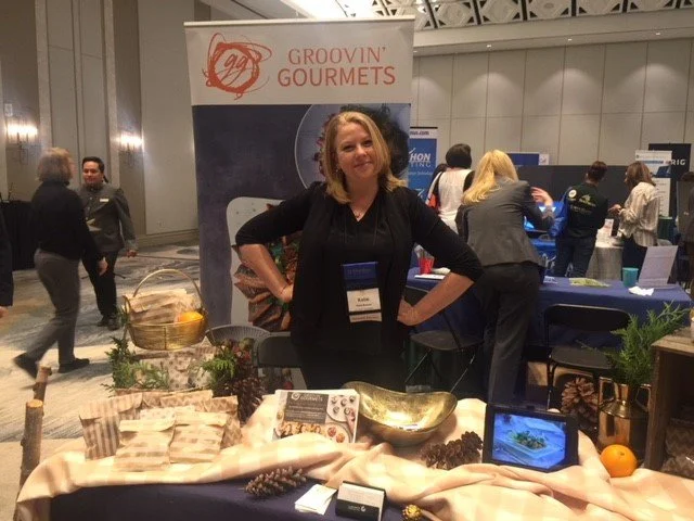 Woman standing behind a display booth with a banner that reads Groovin' Gourmets, showcasing food items and pine cones at a trade show or conference.