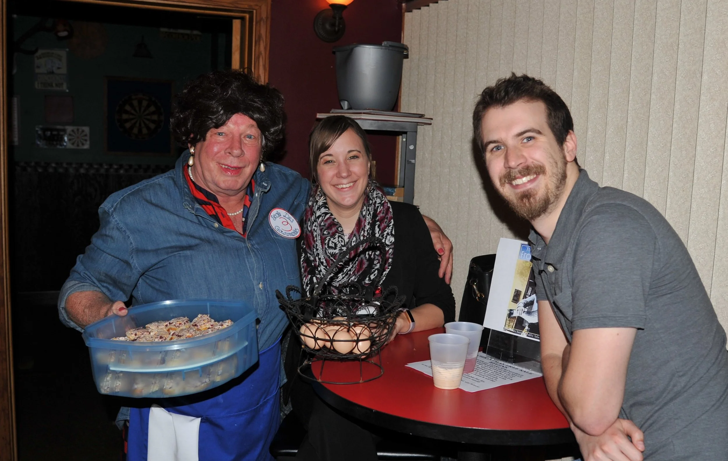 Three people standing behind a red table, smiling at the camera. The person on the left is wearing a black wig, pearl earrings, a blue shirt with a name badge, and holding a blue container of cookies. The person in the middle is wearing a patterned s