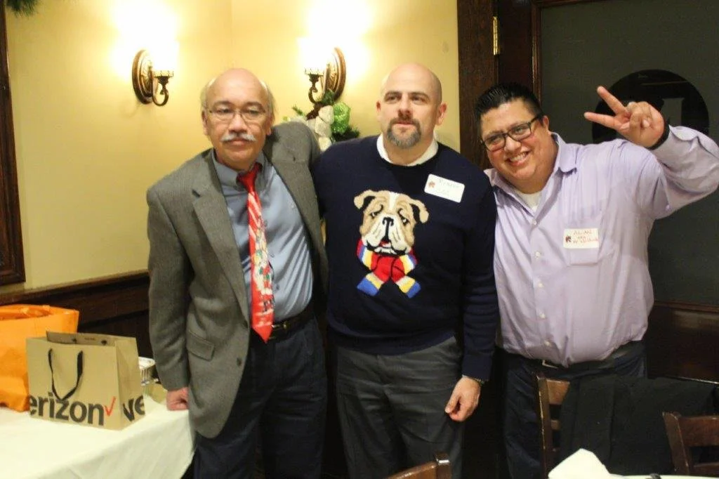 Three men standing together at a social event. The man on the left has gray hair and a mustache, wearing glasses, a gray suit, and a colorful tie. The man in the middle is bald with a beard, wearing a dark sweater with a bulldog motif. The man on the