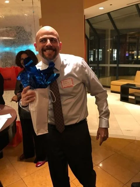 A man with a shaved head and beard smiling, holding a gift bag with blue tissue paper, in a lounge area with large windows and modern furniture.