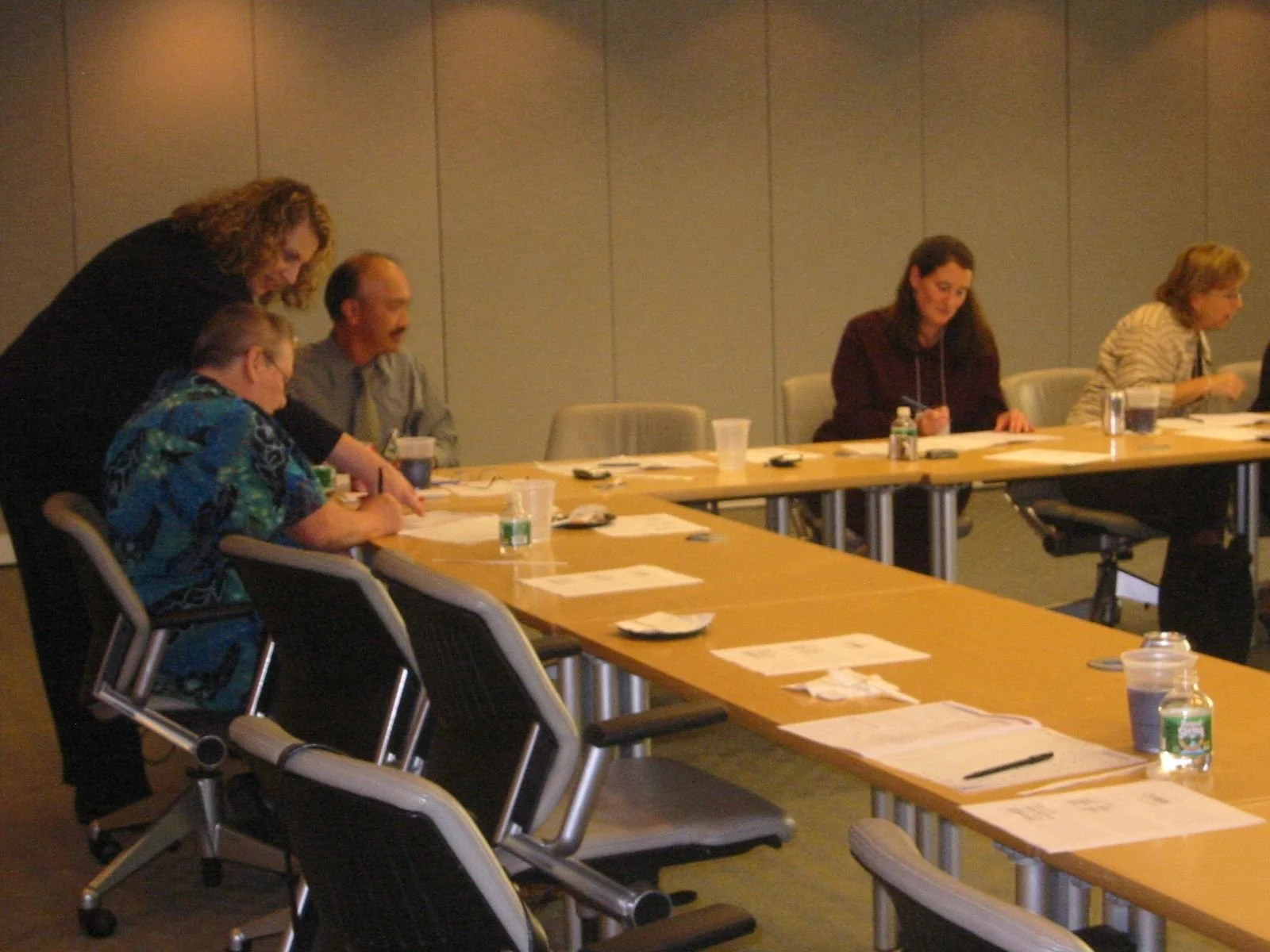A group of six women sitting at a long conference table in a meeting room, engaged in a discussion, with papers, drinks, and personal items on the table.