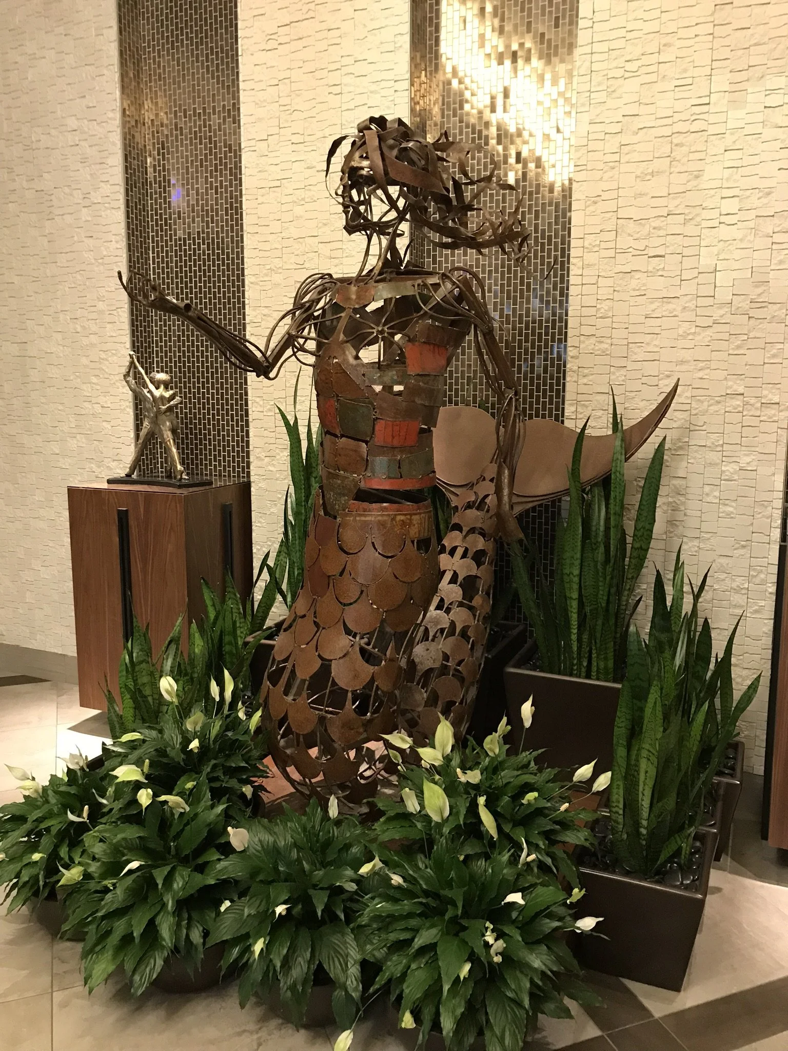 Indoor decorative sculpture of a woman made of metal, surrounded by potted plants with green leaves and white flowers. Background features beige wall with textured panels and a small statue of a dancing figure.
