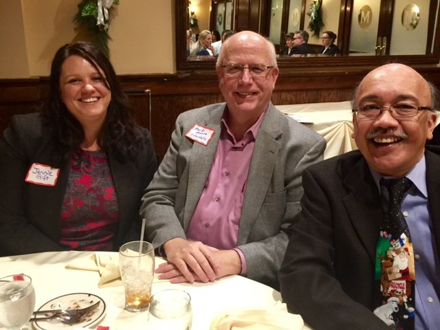 Three people smiling and sitting at a dinner table in a restaurant. The woman on the left has dark hair and is wearing a black blazer with a red top. The man in the middle has glasses, a gray blazer, and a pink shirt. The man on the right has glasses