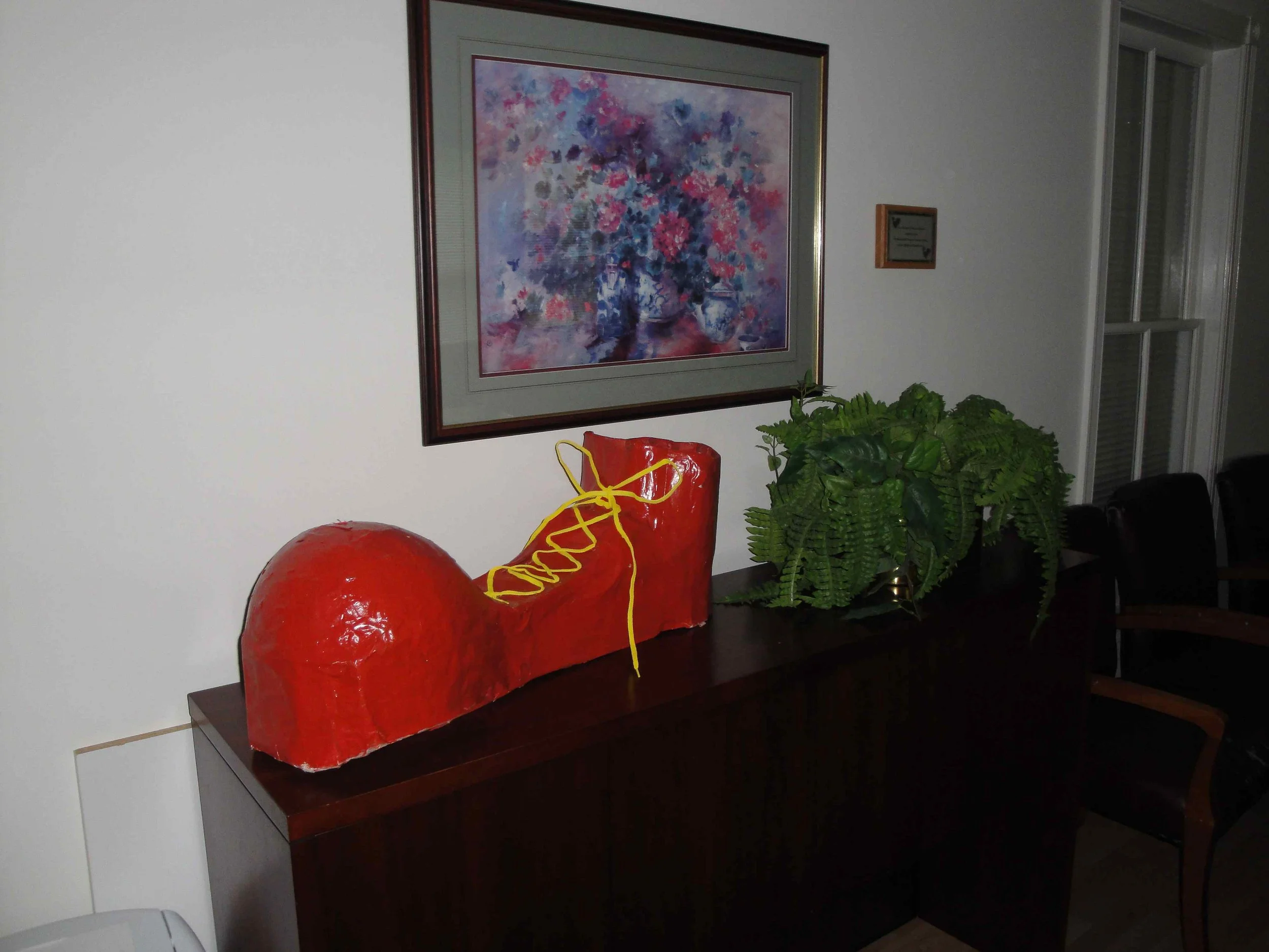 A red sculpture of a vintage car car with yellow attachment. The sculpture is placed on a dark wooden table next to a potted fern plant. There is a large, colorful, abstract floral painting hanging on the white wall behind it.