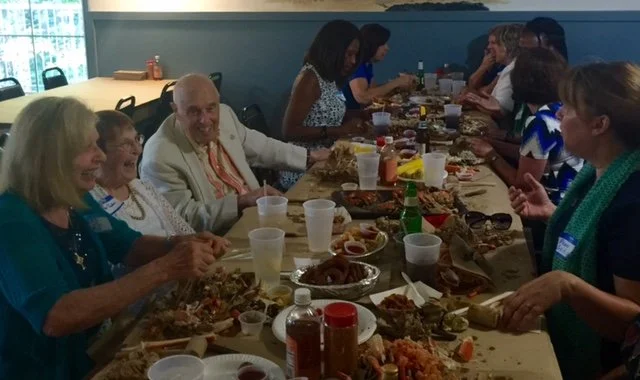 People gathered around a long table enjoying a meal, with drinks and various dishes, in a casual indoor setting.