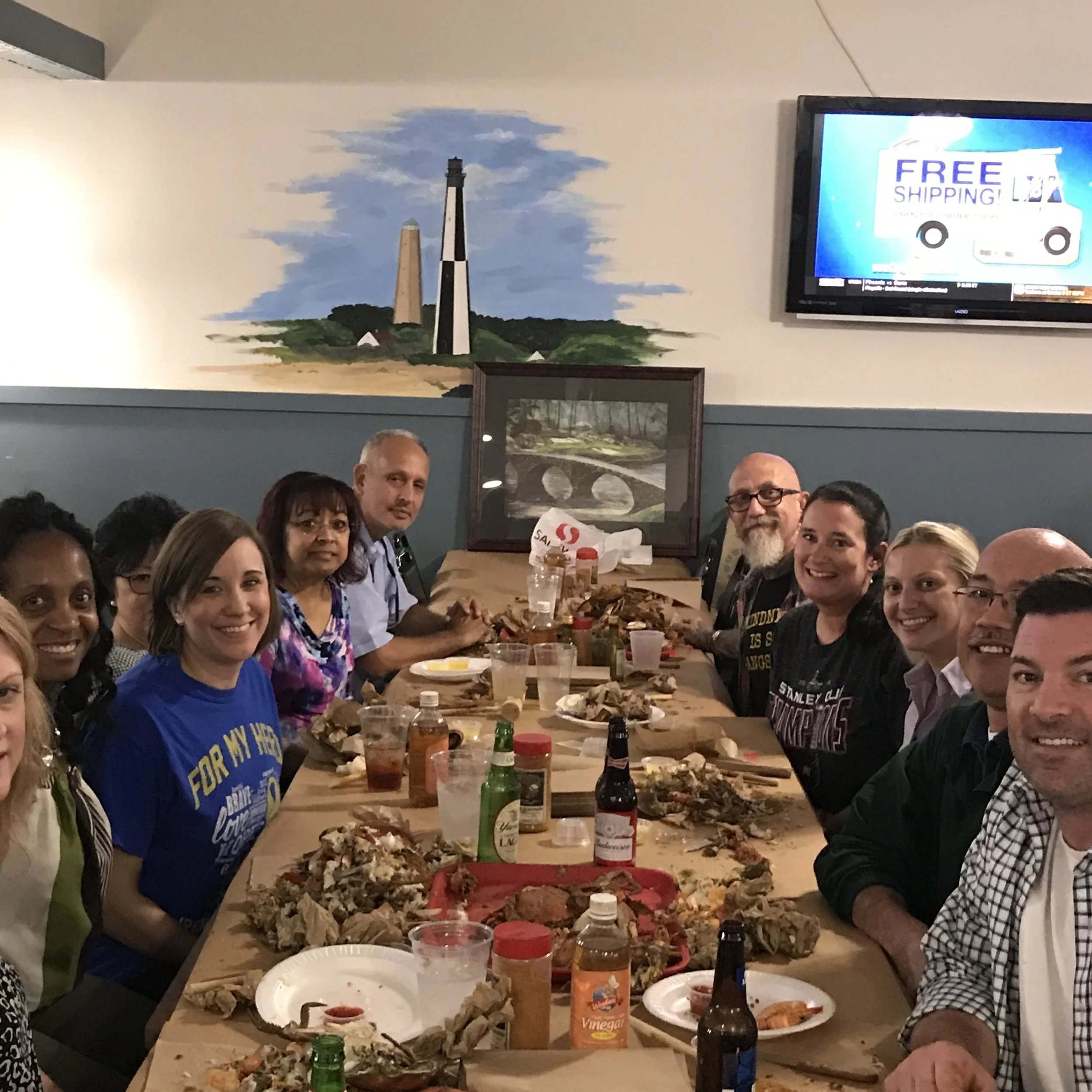 Group of people gathered around a table eating a meal with pizza and drinks. A mural of two lighthouse towers and a partly cloudy sky is on the wall in the background. A TV screen displays an advertisement for free shipping with a delivery truck grap