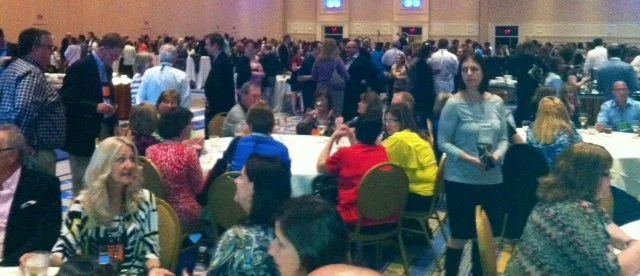 Large crowded event hall with many people seated at round tables and standing, engaged in conversations, with some holding drinks.