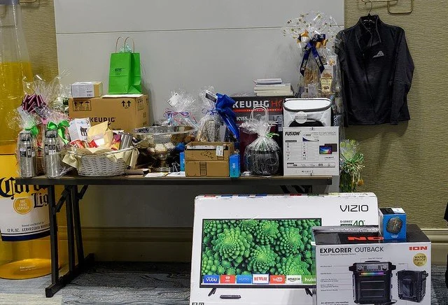 Table filled with gift baskets, boxes, and various items; a green gift bag in the background; a black jacket hanging on the wall; a flat-screen TV and a printer in front of the table; some electronic devices and packages on the right.