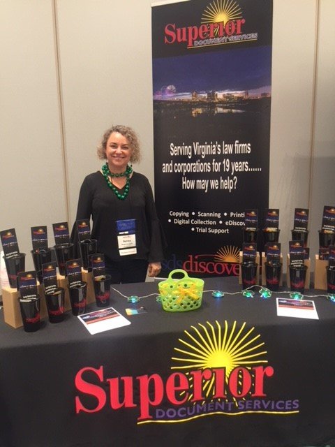 A woman standing behind a table at a trade show booth for Superior Document Services, with a banner in the background. The table displays multiple copies of the company's brochure, a yellow basket, and some small LED lights.