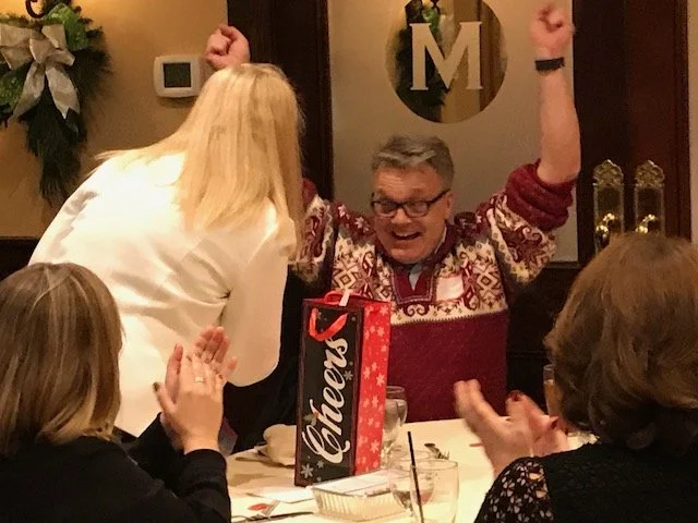 A man celebrating with raised arms at a dinner table surrounded by women clapping and smiling, with a gift bag labeled "Cheers" on the table.
