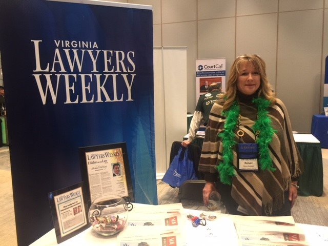 Woman standing at Virginia Lawyers Weekly booth during a conference, wearing a green feather boa and name badge, with informational materials and a fishbowl on the table.
