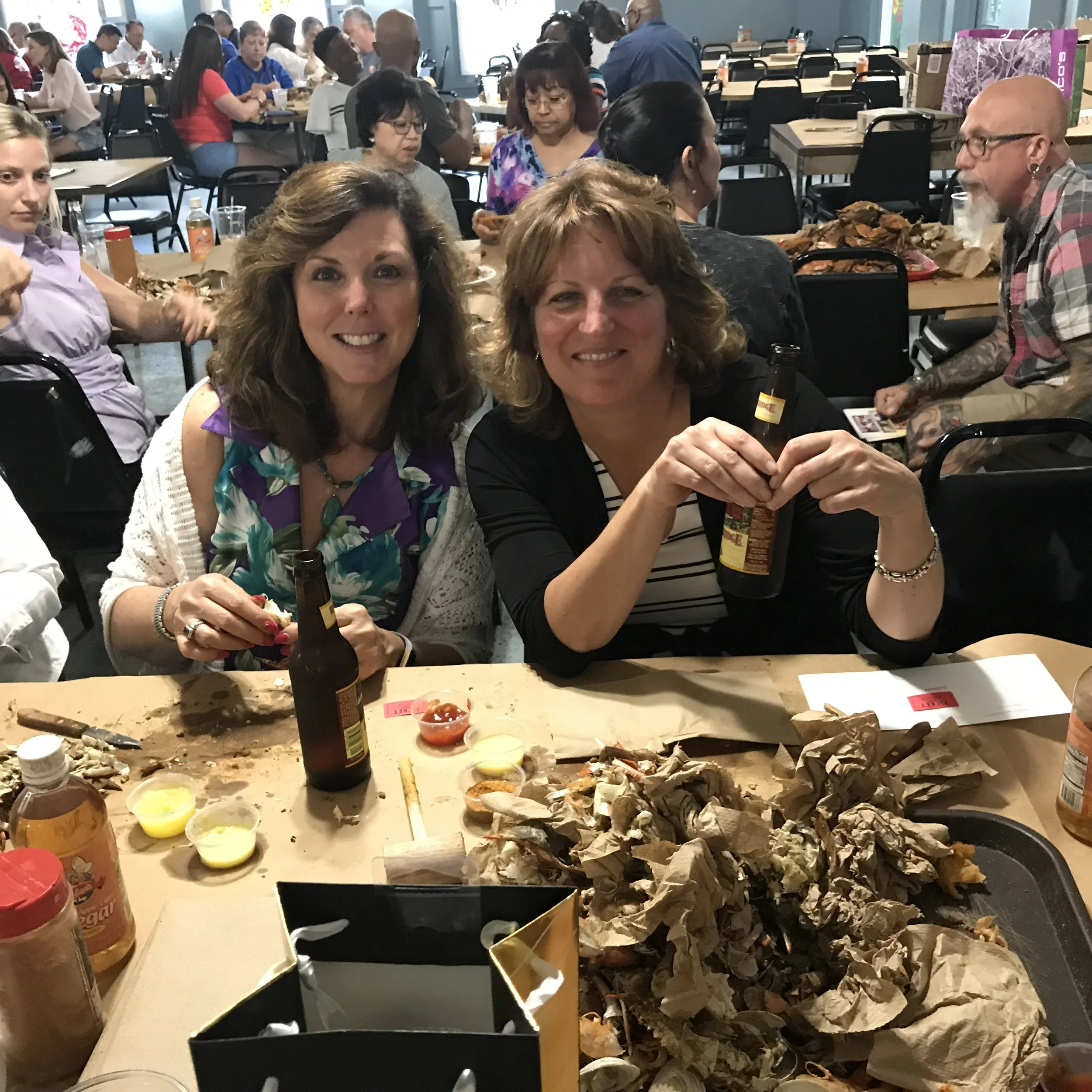 People sitting at tables in a crowded room, eating and socializing, with a table in the foreground covered in trash, shells, and condiments, indicating a casual or celebration event.
