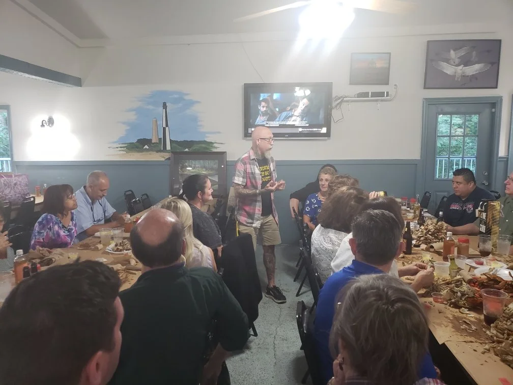A man with glasses, a beard, and tattoos wearing a plaid shirt and shorts is speaking to a group at a restaurant or banquet hall during a casual gathering or party. The group is seated at tables with food and drinks, and some are taking photos or lis