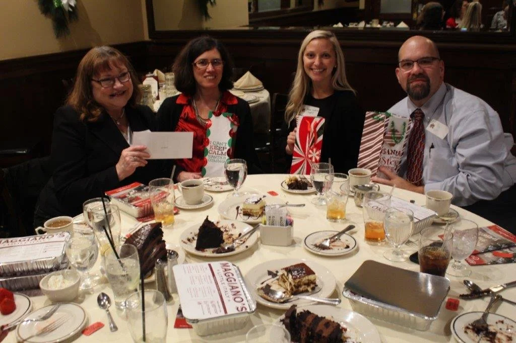 Four people sitting at a round table with dessert, drinks, and gift bags, celebrating in a restaurant.