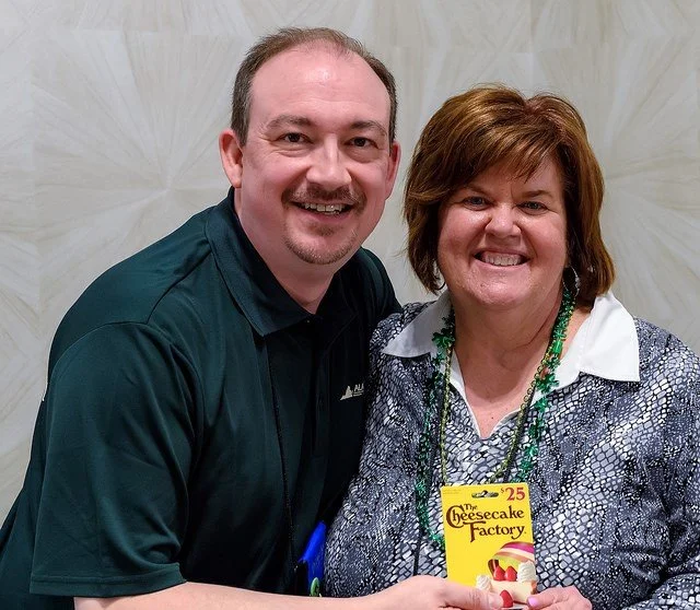 Two smiling people, a man and a woman, posing together. The woman is holding a small Cheesecake Factory box or booklet.
