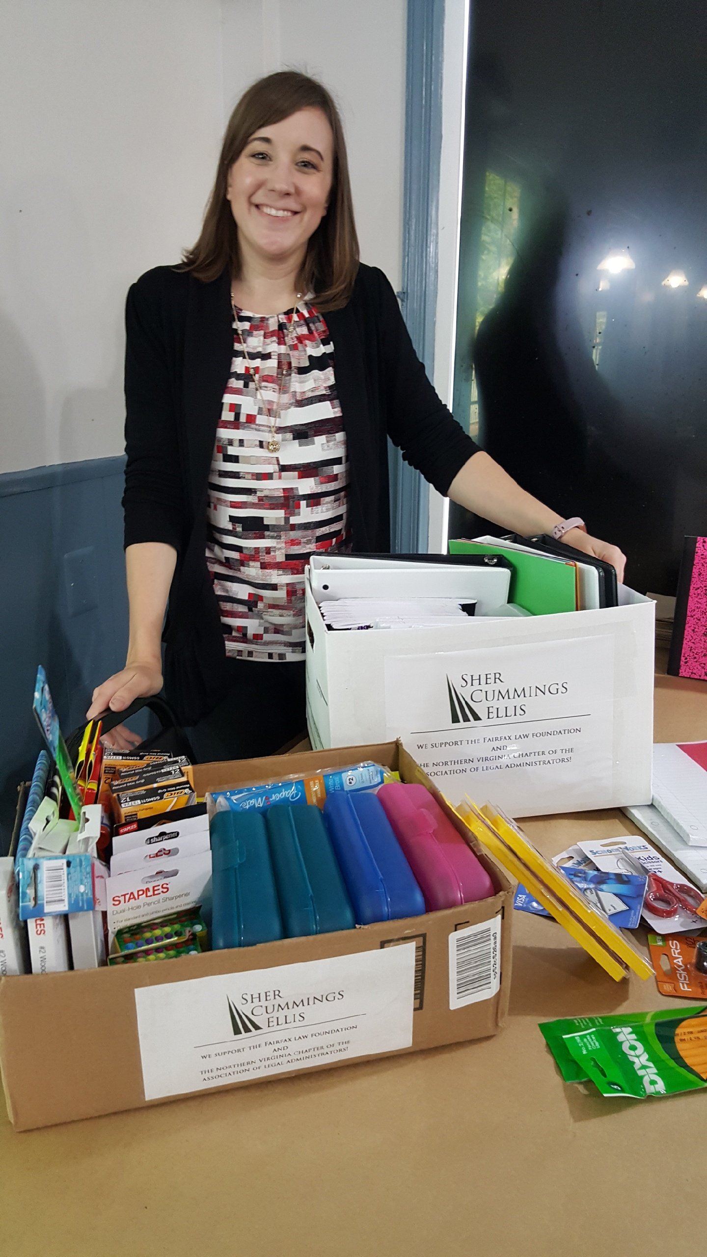 A woman with shoulder-length hair smiling at the camera. She is standing behind organizing supplies on a table, which includes pens, scissors, and various stationery items. There are boxes labeled with "Sher Cummings Ellis" supporting a foundation. T