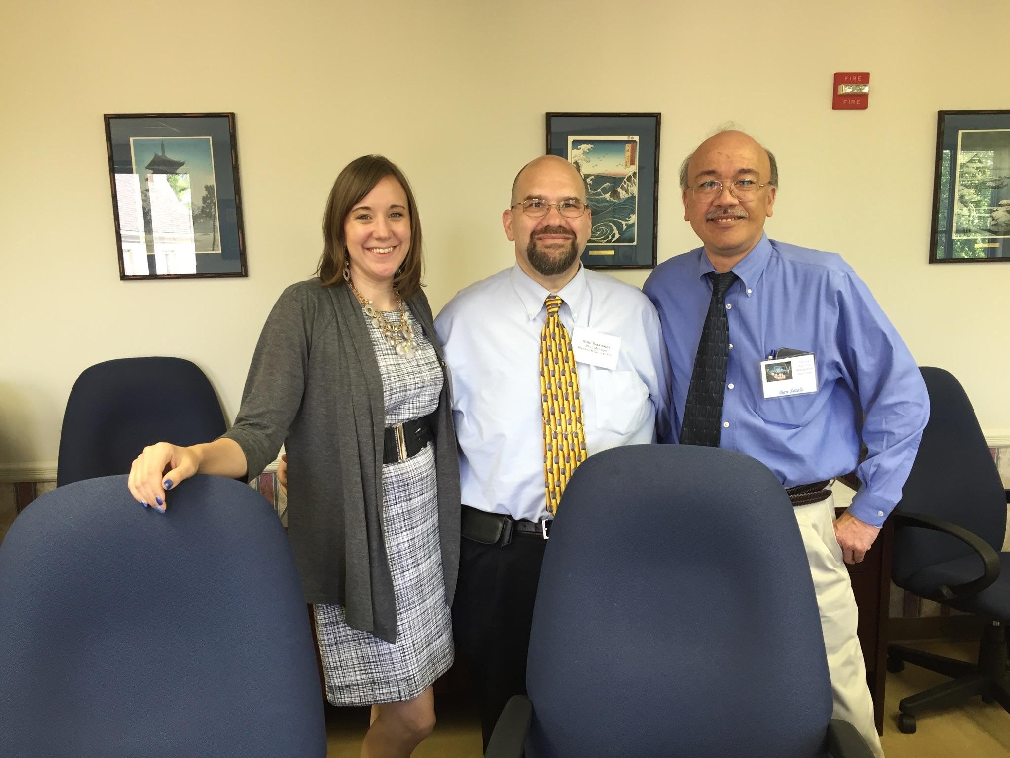 Three people standing together in a meeting room, smiling at the camera. The woman on the left has shoulder-length brown hair and is wearing a gray blazer and plaid dress. The man in the middle has a beard, glasses, and is wearing a light blue shirt 