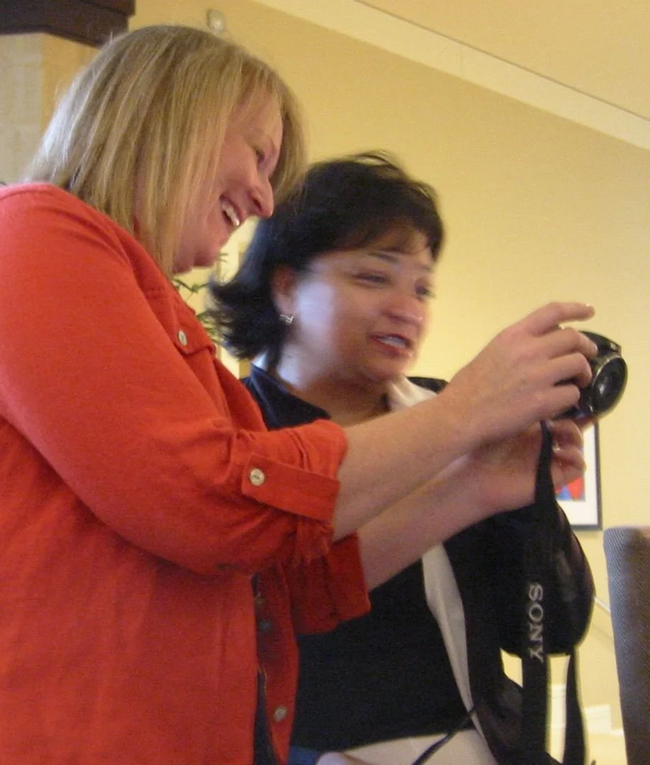 Two women are smiling and looking at a camera, with one holding a Sony camera and camera bag.