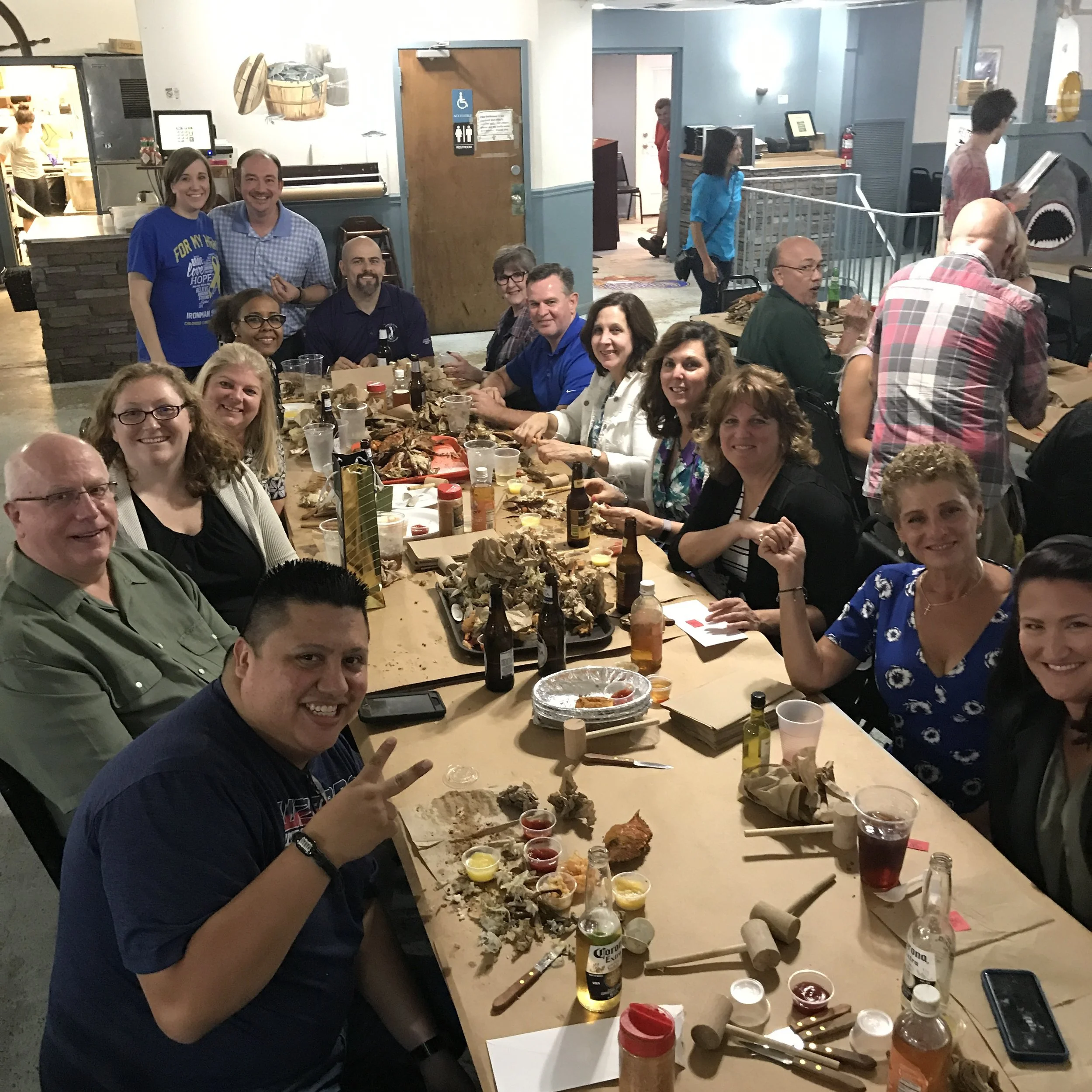People gathered around a table at a restaurant, enjoying a meal with various dipping sauces, beer bottles, and food scraps, with some individuals smiling and making peace signs.