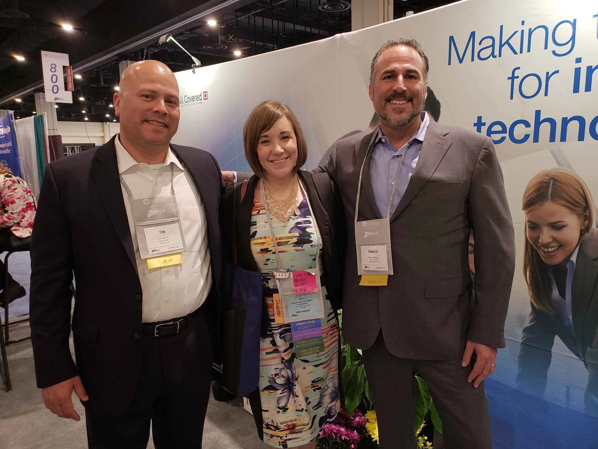 Three people standing together at an indoor event, with a backdrop that has text about making a difference for technology and a company logo. They are smiling, wearing name tags, and are dressed in formal attire; two men in suits and a woman in a col