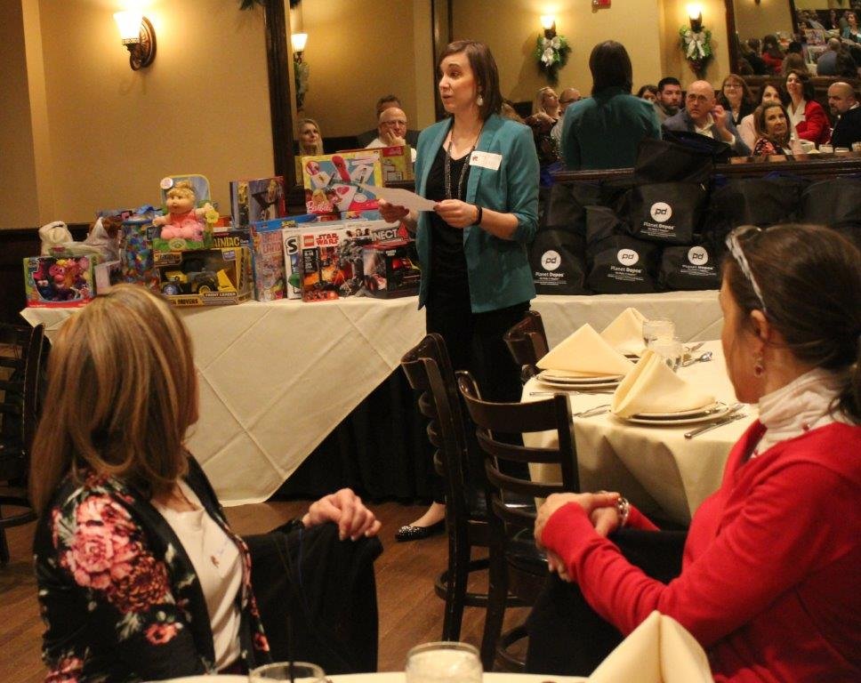 A woman in a teal blazer and black pants stands at a dining table speaking to two seated women at a banquet. The table has folded yellow napkins, utensils, and a display of toys, including dolls and action figures. In the background, a large group of