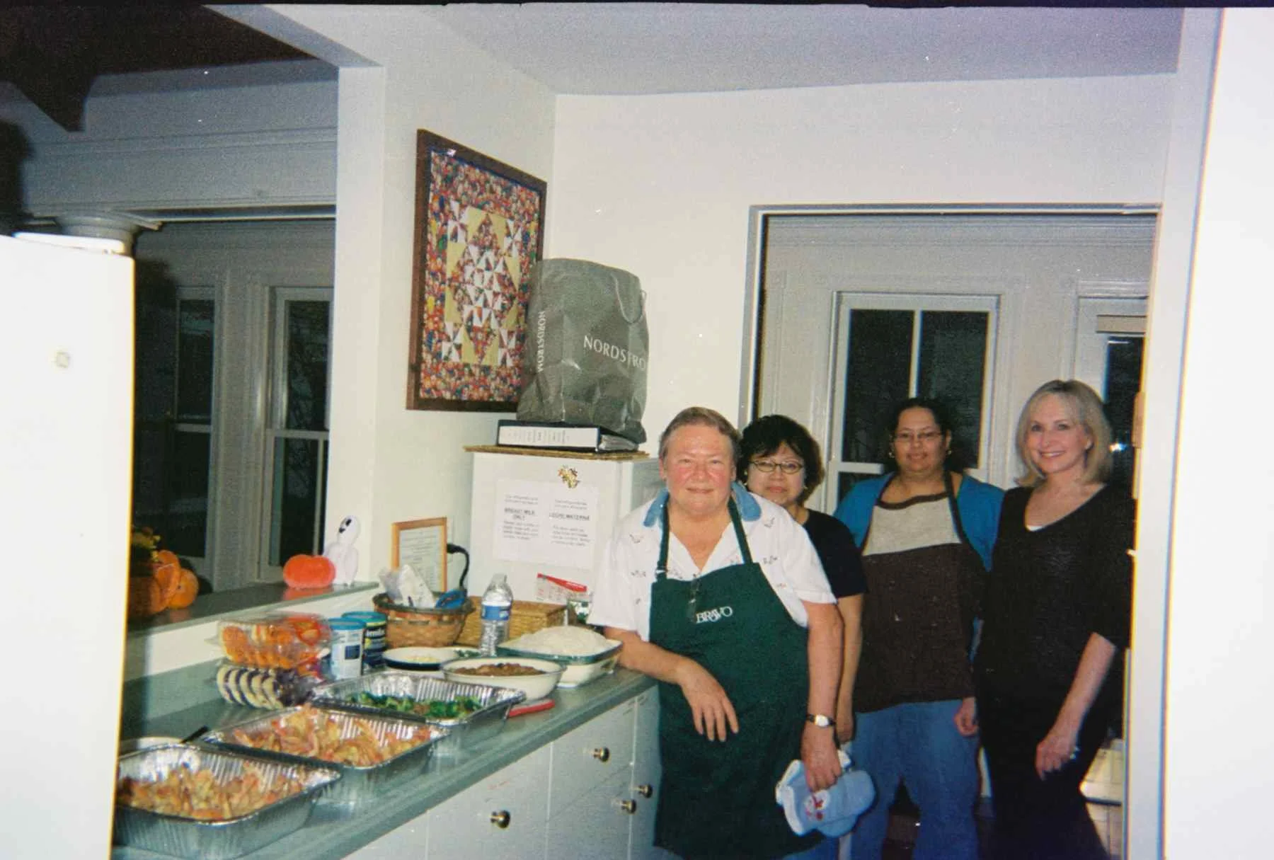 Four women stand in a kitchen behind a counter with dishes of food, including rolls and baked items, and various food items. The woman in front wears a white shirt and green apron, smiling. The other women wear casual clothes and glasses. There are w