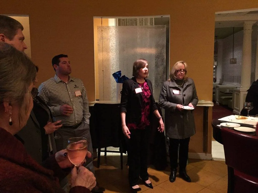 People gathered in a dimly lit room, with a woman speaking or presenting, others holding drinks, likely at a social or professional event.