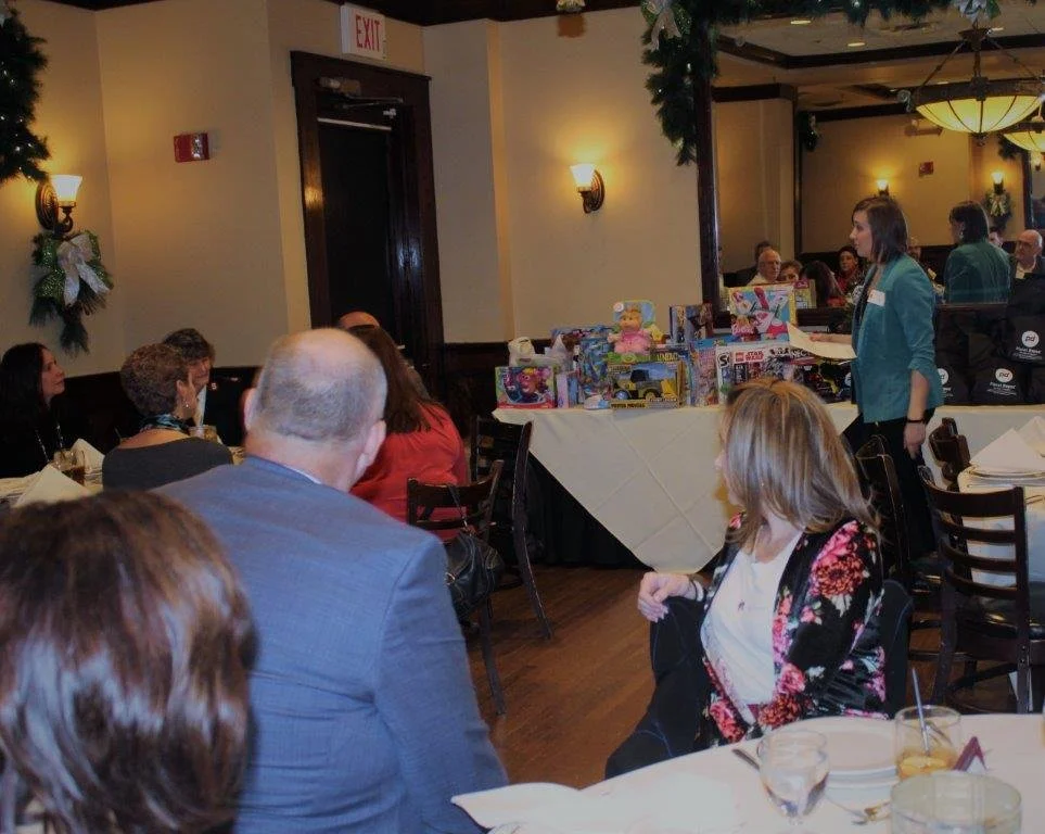 Indoor event at a restaurant or banquet hall with seated guests, a table with toys or gifts, and a woman standing near the toys, possibly speaking or presenting.