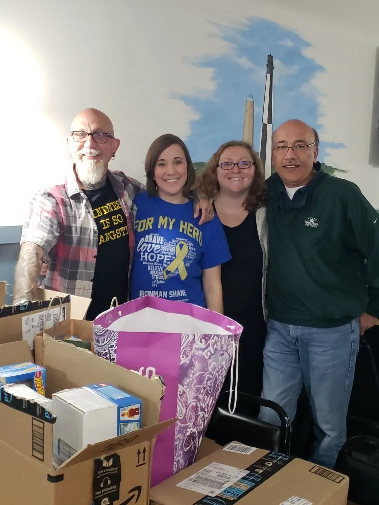 Four smiling people standing together, with boxes and a donation bag in the foreground, indoors with a mural of a blue sky and what appears to be a lighthouse in the background.