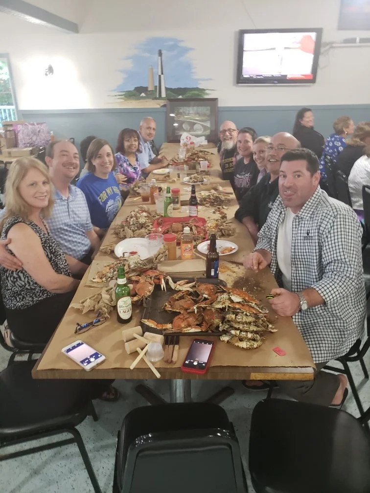 A group of people sitting around a long table enjoying a seafood feast with lobster and crab, with drinks and condiments, in a restaurant setting.