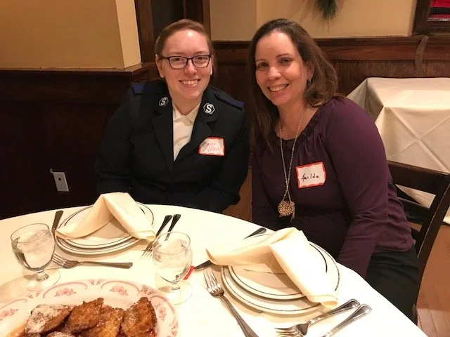 Two women sitting at a dining table with white napkins, plates, glasses, and silverware. One woman is wearing a dark uniform with a badge, and the other is wearing a purple top with a necklace. They are smiling and posing for the photo in a restauran