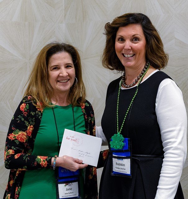 Two women smiling for the camera, one holding a card, standing against a beige wall. Both wearing lanyards with badges, one with a green shamrock necklace.