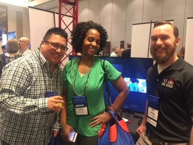 Three people smiling at a conference, standing in front of a TV screen, wearing name badges, with other attendees in the background.