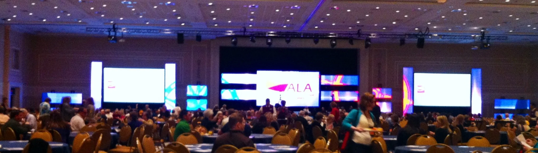 A large conference room filled with many people sitting at tables, facing a stage with multiple large screens displaying a presentation. The ceiling is decorated with small, evenly spaced lights.