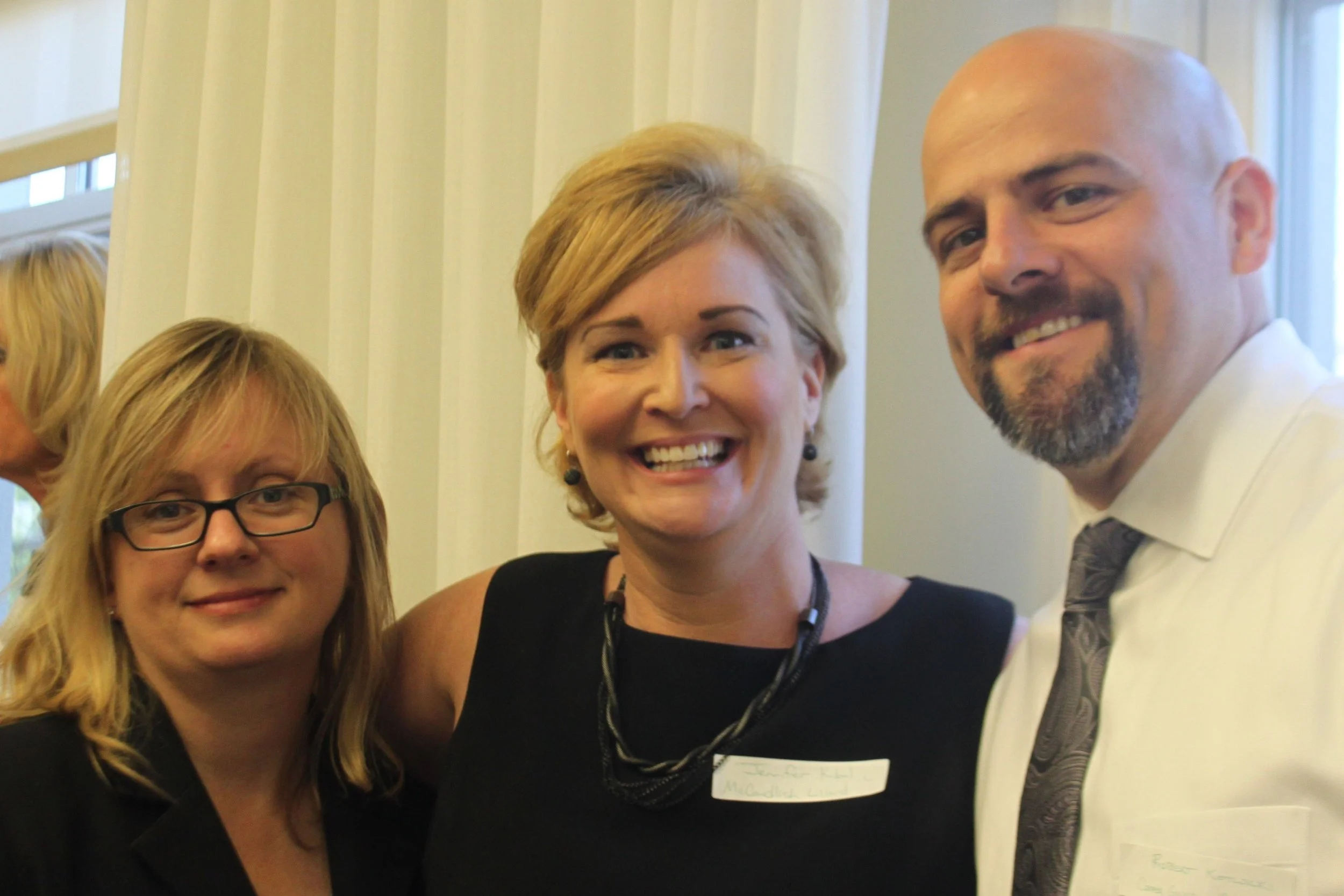Four people, two women and one man, posing for a photo indoors in front of a cream-colored wall with vertical panels. All are smiling and looking at the camera. The woman on the left wears glasses and has blonde hair, the woman in the middle has shor