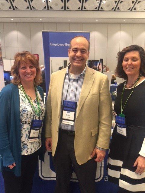 Three people smiling at a conference, standing in front of a booth with a sign that says 'Employee Ben...'