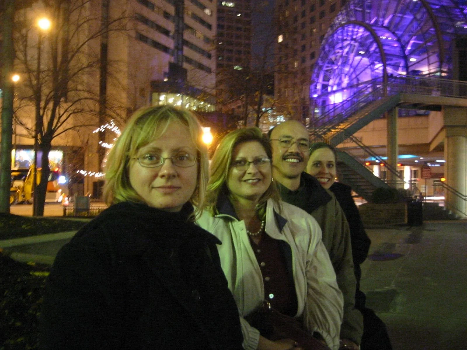 Four people posing outdoors at night in an urban setting, with a lit building and a glass-enclosed staircase in the background.