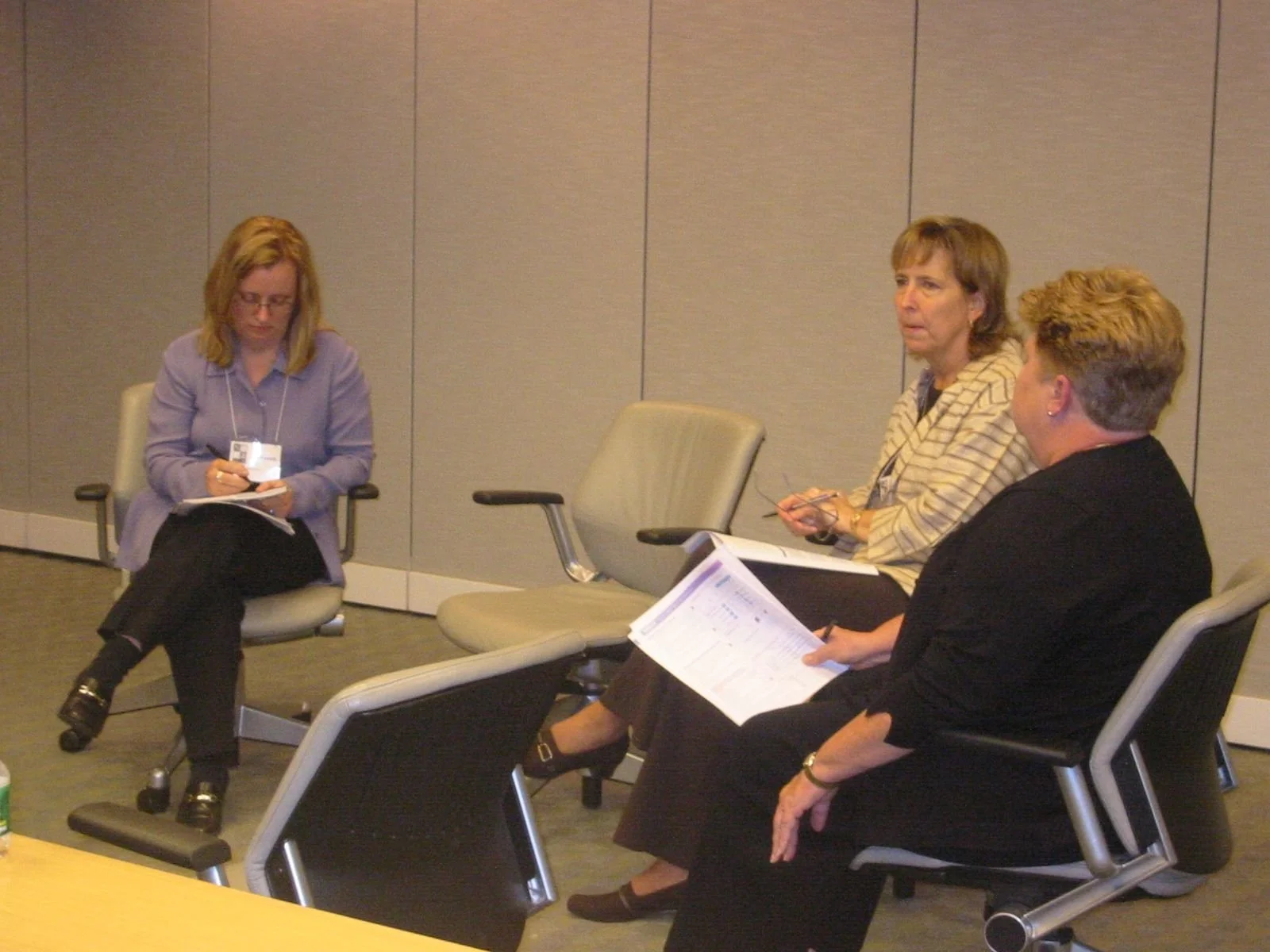 Three women sitting in chairs in a conference room, engaged in a discussion or interview, with one woman taking notes.