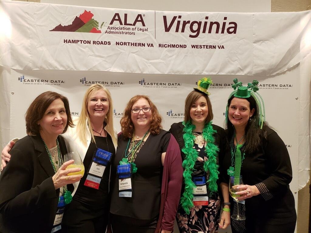 Five women at a Virginia ALA event, wearing green accessories, some with leprechaun hats, smiling and holding drinks, standing in front of a banner and backdrop.