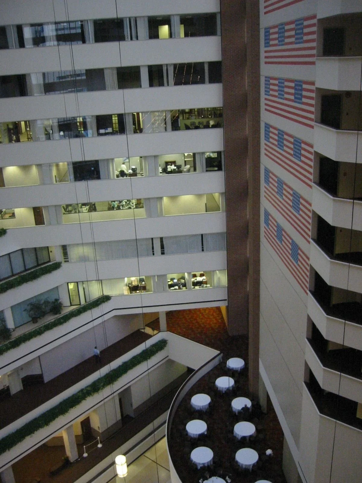 Interior view of a multi-story office building with multiple floors visible. The building features glass walls, office spaces, and a large American flag mural on one wall. There is a circular area with tables and chairs at the bottom of the image, an